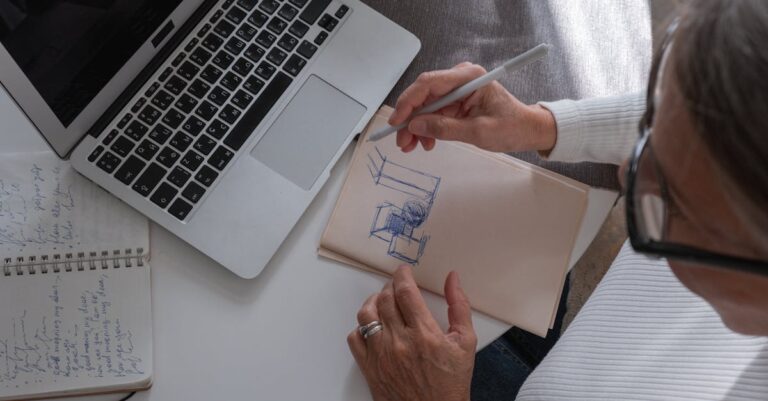 Mãos anotando variações de título em caderno com post-its e laptop por perto, vista superior