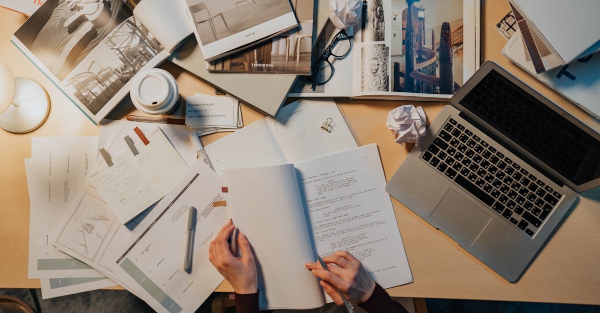 Mesa de trabalho com laptop, rascunhos, caderno e xícara de café vistos de cima, sugerindo organização da escrita