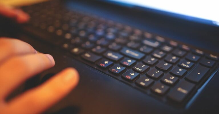 Mãos registrando anotações e logs em caderno ao lado de laptop com tela desfocada
