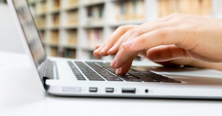 Laptop aberto sobre mesa de biblioteca com prateleiras ao fundo e mãos no teclado