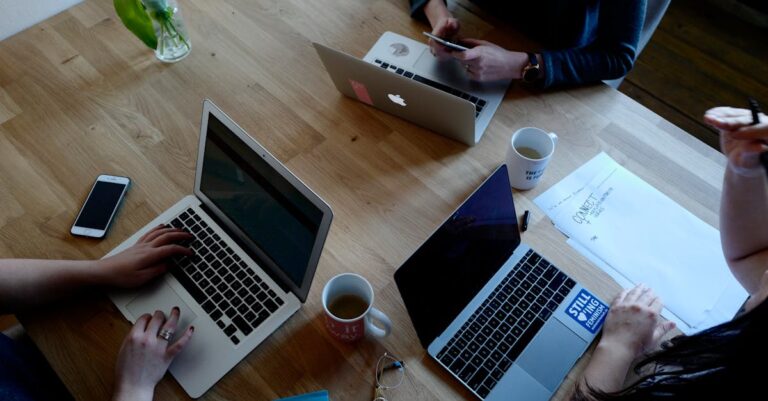 Mãos de participantes digitando em laptops e rascunhos, cronômetro sobre a mesa, sessão de escrita coletiva.