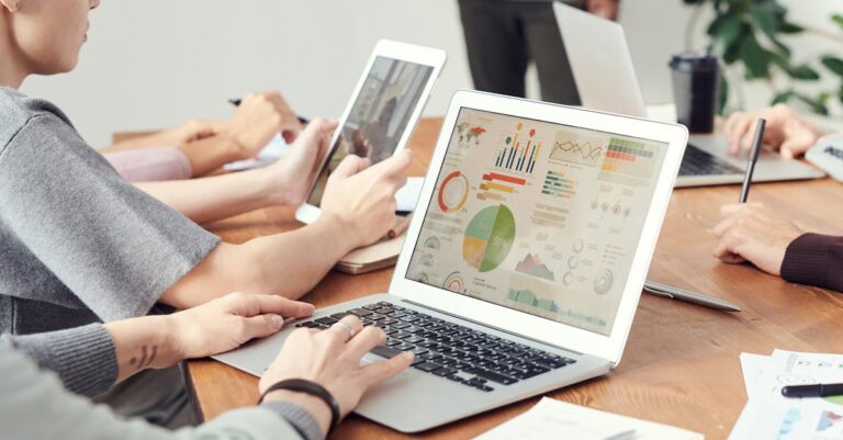Tela com gráficos e tabelas impressas sobre mesa, notebooks e anotações, indicando apresentação de dados