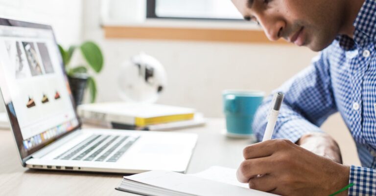 Mãos escrevendo em diário curto ao lado de laptop e caneca, registrando progresso semanal.