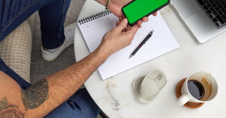 Mãos segurando um celular com tela de contato de emergência, bloco de notas e caneta sobre a mesa.