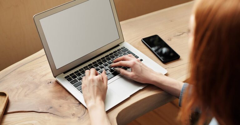Mãos digitando em laptop com projeto de uma página ao lado, ambiente de estudo organizado