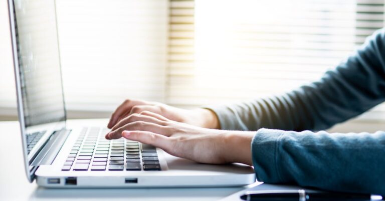 Mãos digitando e PDFs digitalizados ao lado do laptop em mesa de escritório.