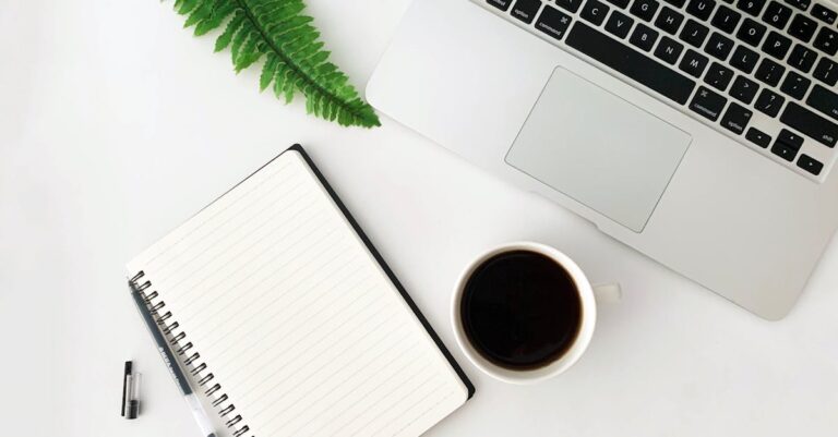 Mesa com laptop aberto, folhas amassadas e caderno com caneta, sugerindo tentativa e erro