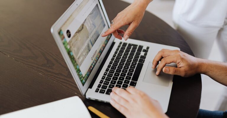 Mãos apontando para rascunhos de pré‑projeto sobre a mesa, materiais de revisão e laptop