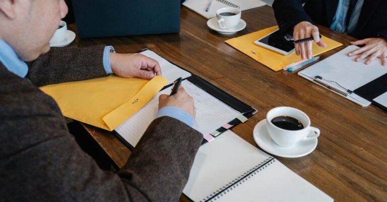 Duas mãos apontando para uma agenda e notas de reunião sobre a mesa, laptop ao lado.