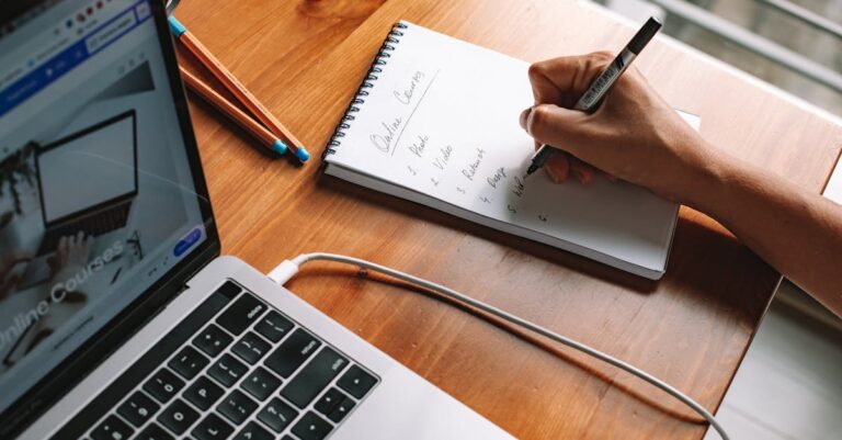 Mãos indicando tela de laptop com interface de base de dados acadêmica, bloco de notas ao lado