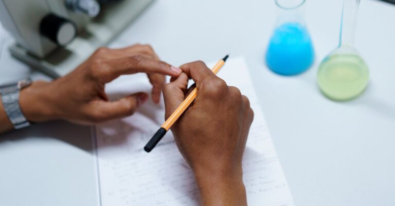 Mãos escrevendo o pré-projeto ao lado de laptop e anotações em ambiente de estudo