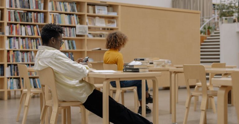 Mesa de biblioteca universitária com documentos de política, laptop e estantes ao fundo