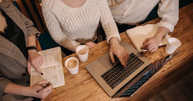 Mãos de equipe colaborando sobre mesa com laptops e post-its, representando divisão de tarefas e atribuições
