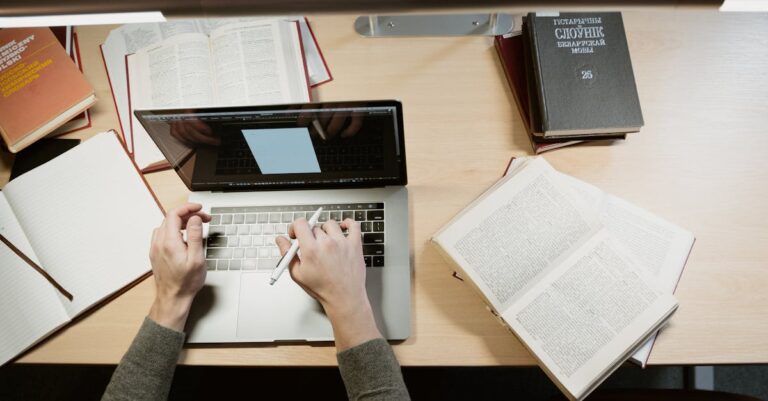 Mãos digitando em laptop com notas e livros abertos, foco na redação da contribuição científica