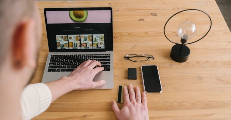 Mãos consultando site institucional da universidade no laptop, smartphone e edital impresso ao lado.