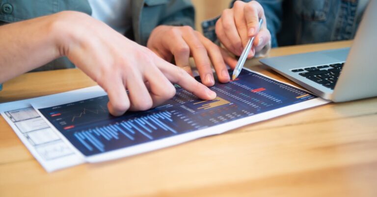 Duas pessoas revisando um resumo/proposta sobre a mesa com laptop e notas espalhadas.