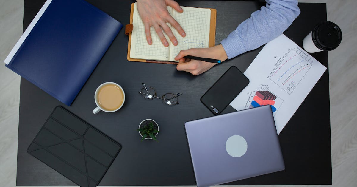 Mesa de estudos com laptop, caderno aberto, caneta e mãos escrevendo sob luz natural
