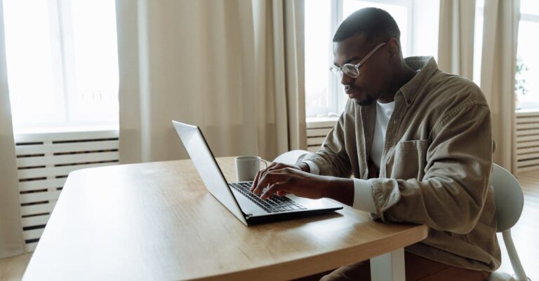 Mãos digitando em laptop com projeto impresso e caneta ao lado, mostrando preparo de proposta para inscrição