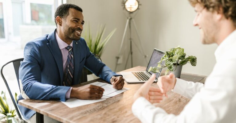 Mesa de escritório com laptop e folhas, duas pessoas discutindo documento, foco nas mãos