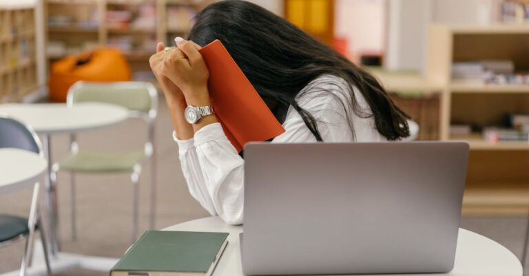 Mãos junto ao rosto sobre a mesa com laptop e caderno, sugerindo exaustão e pressão acadêmica.