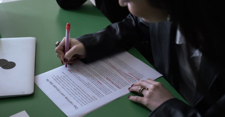 Mãos revisando rascunho de proposta com caneta vermelha e laptop ao lado