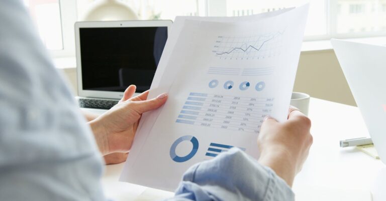 Mãos de avaliadores analisando currículos e projetos sobre mesa, documentos e laptops visíveis