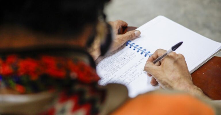 Estudante pensativo escrevendo notas em caderno em mesa clara, com expressão concentrada e iluminação natural.