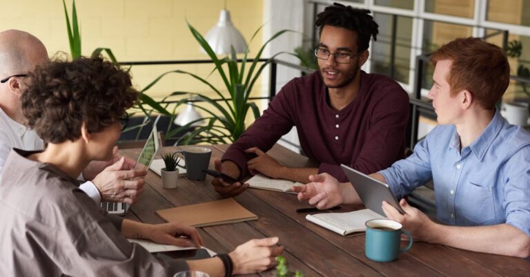 Grupo de pesquisadores discutindo em reunião com laptop e papéis em mesa iluminada naturalmente