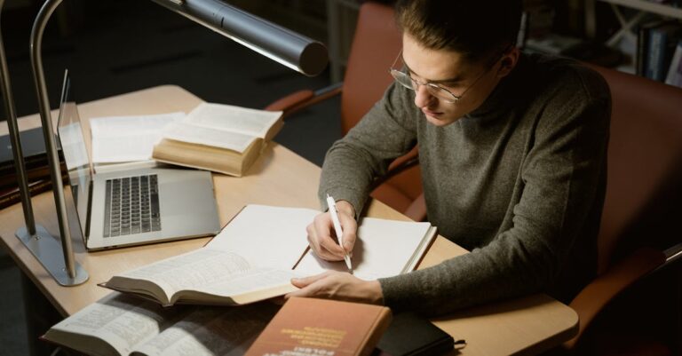 Estudante pesquisador digitando notas em laptop com pilha de artigos acadêmicos ao lado em mesa organizada