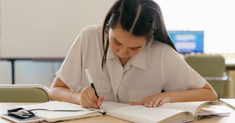 Mulher escrevendo notas acadêmicas em caderno com laptop ao lado em ambiente profissional iluminado